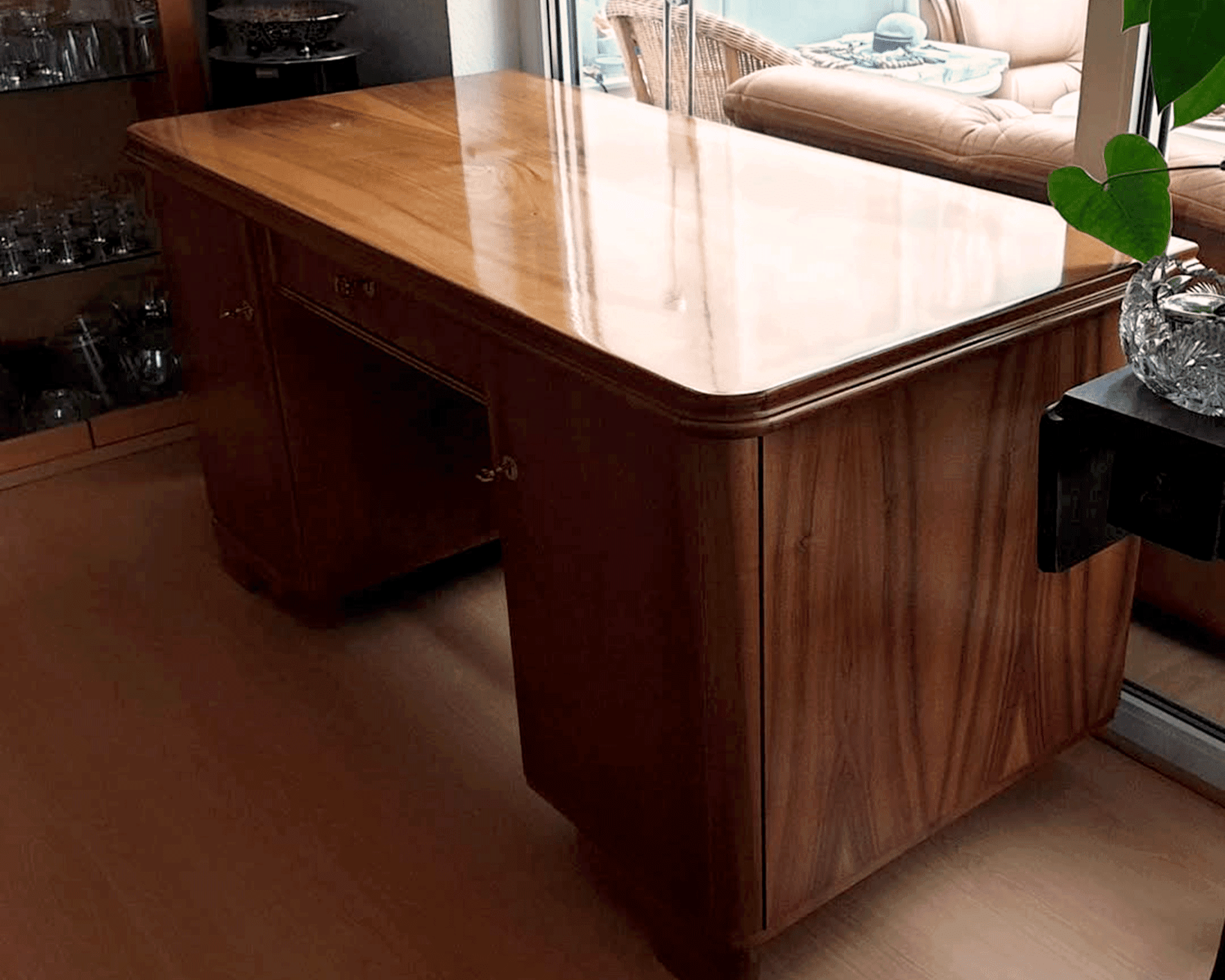 Ein eleganter Holzschreibtisch mit glänzender Oberfläche, der in der Nähe eines Fensters steht. Eine Kristallvase mit grünen Blättern steht auf einem Regal in der Nähe und schafft ein warmes, einladendes Ambiente.